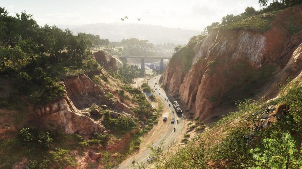 GTA 6'da yeşil ağaçlar ve dik yamaçlar arasından kıvrılarak uzanan bir dağ yolu ve uzaktaki köprü manzarası